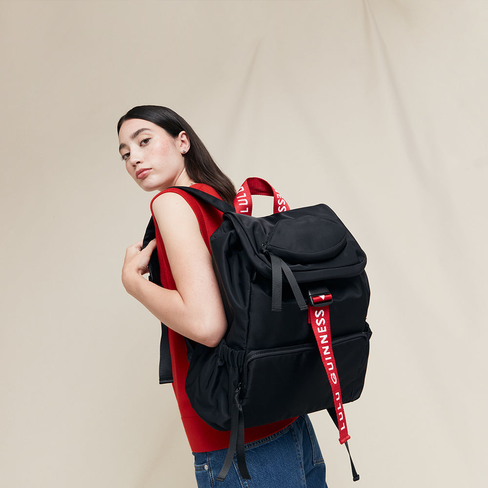 Person wearing a black backpack with 'Lulu Guinness' branded red straps on a beige background