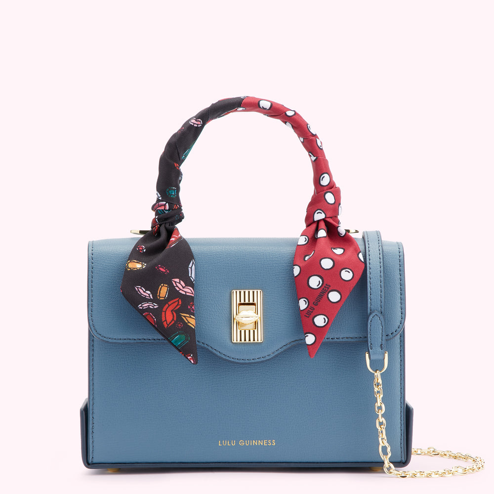 A teal blue Lulu Guinness satchel with a gold clasp and chain strap, adorned with a colorful silk scarf tied around the handle.