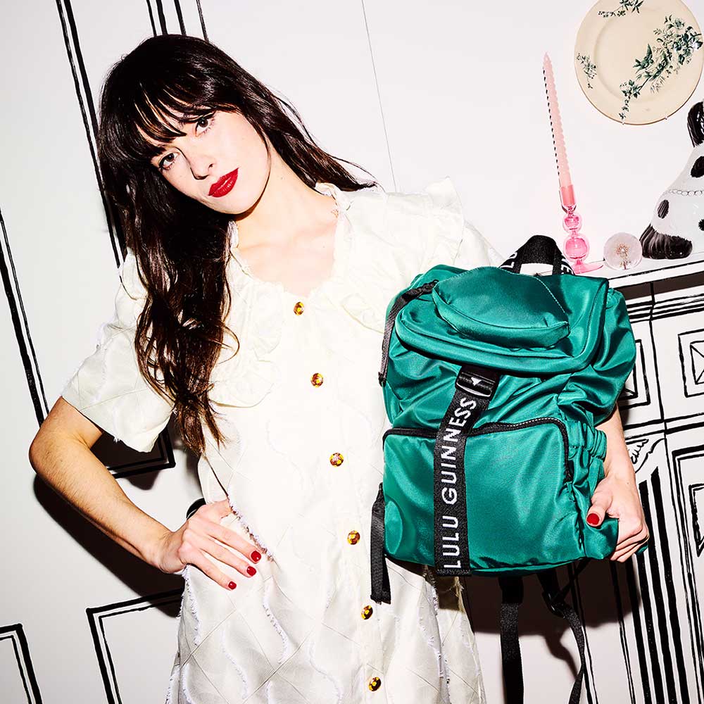 Model with brown hair wearing a white dress and pink lipstick holding a emerald green nylon backpack with branded 'Lulu Guinness' straps.