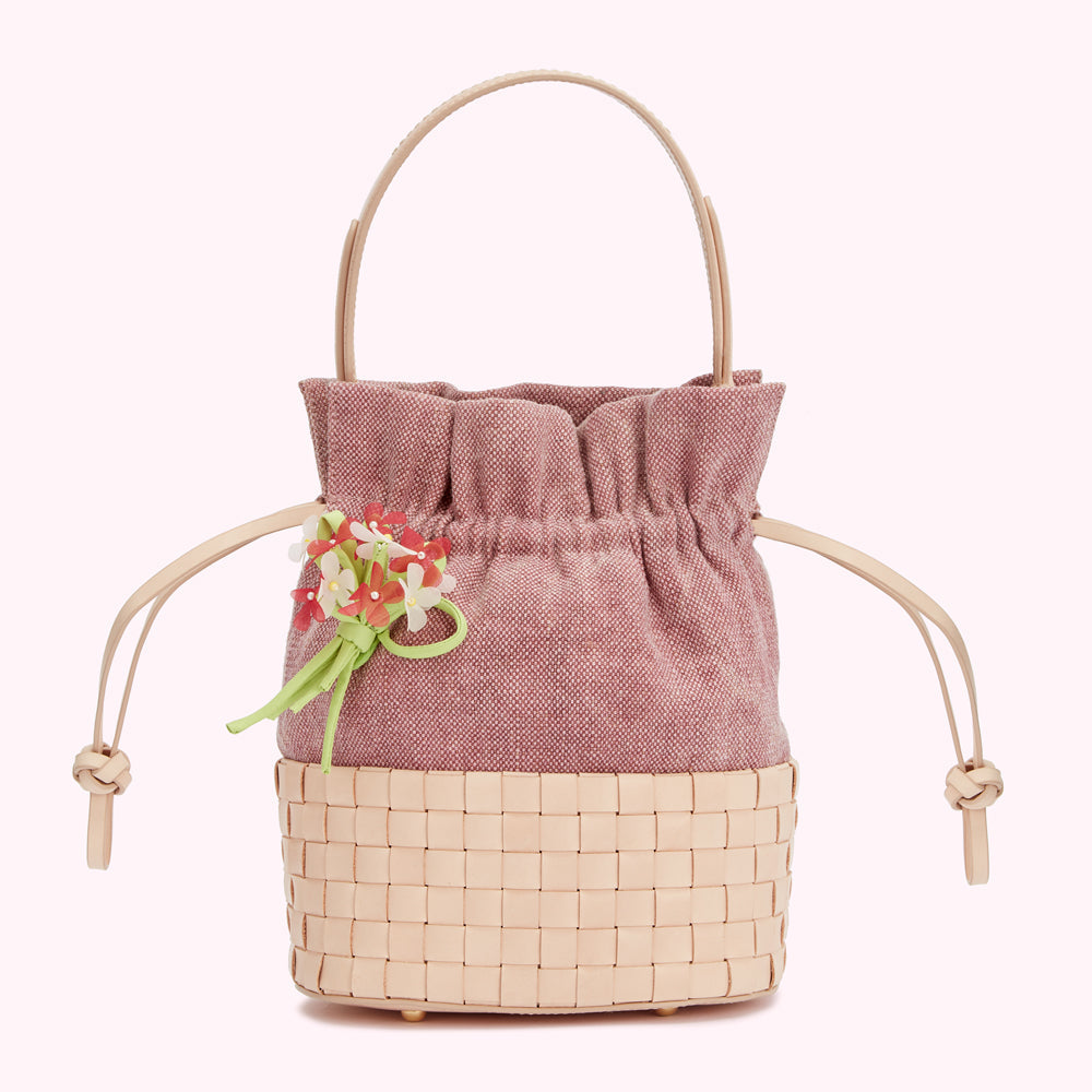 Dusty rose and beige woven bucket bag with a leather top handle and drawstring closure, accented with a small fabric flower bouquet.