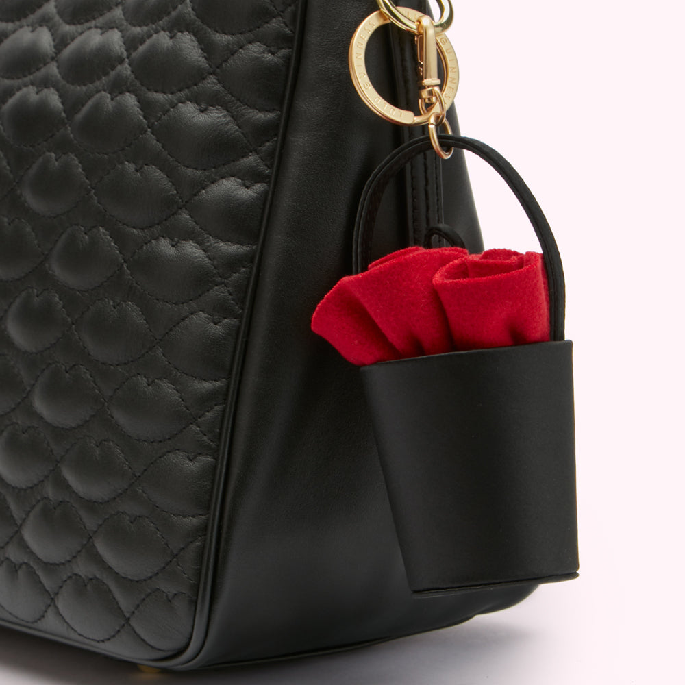 Close-up of a black quilted leather handbag with a red fabric flower detail attached to a small black bucket-shaped accessory hanging from the bag's handle.