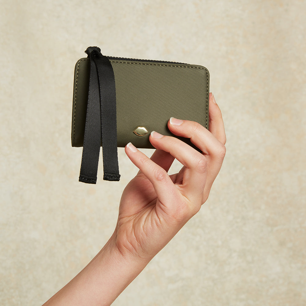 Hand holding a green wallet with a black strap against a beige background