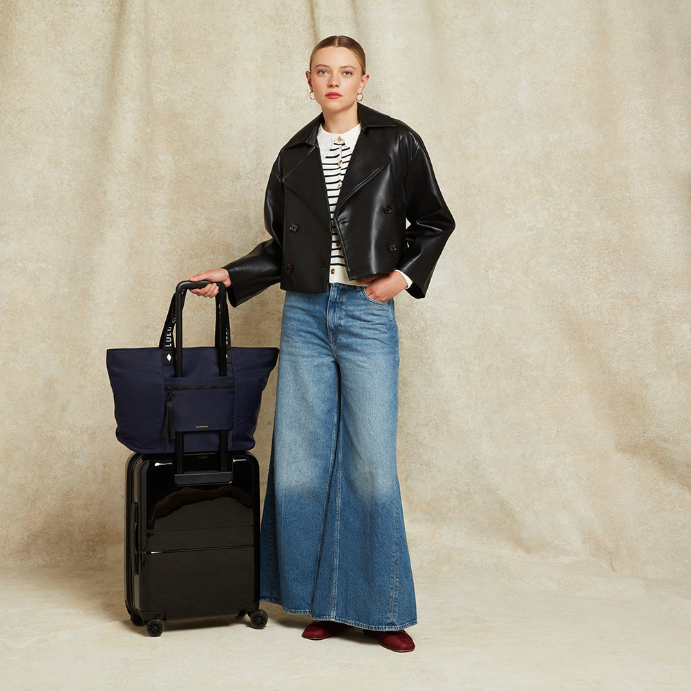 Woman in black leather jacket and blue jeans holding a navy bag slot on top of a black suitcase against a beige background