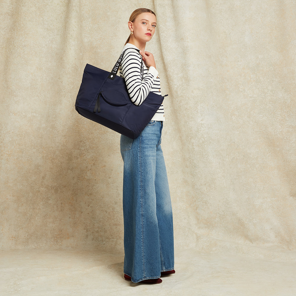 Women in a striped top and jeans carrying a navy blue tote bag on her shoulder against a beige background
