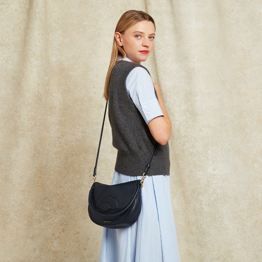 Woman carrying a navy leather handbag showing the crossbody strap against a beige curtain background