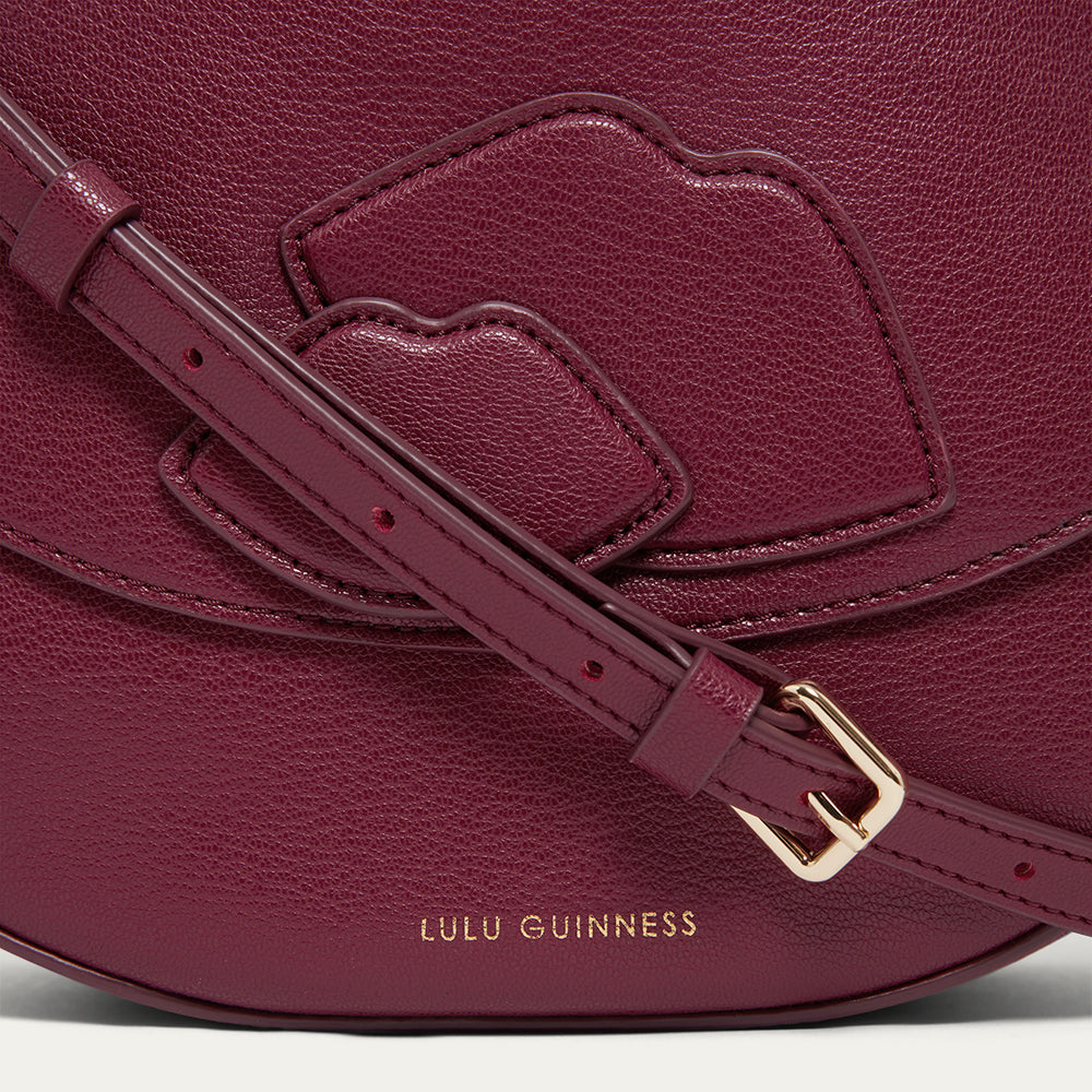 Close-up the lip detail and gold buckle of a burgundy leather handbag with gold printed ' Lulu Guinness' branding.