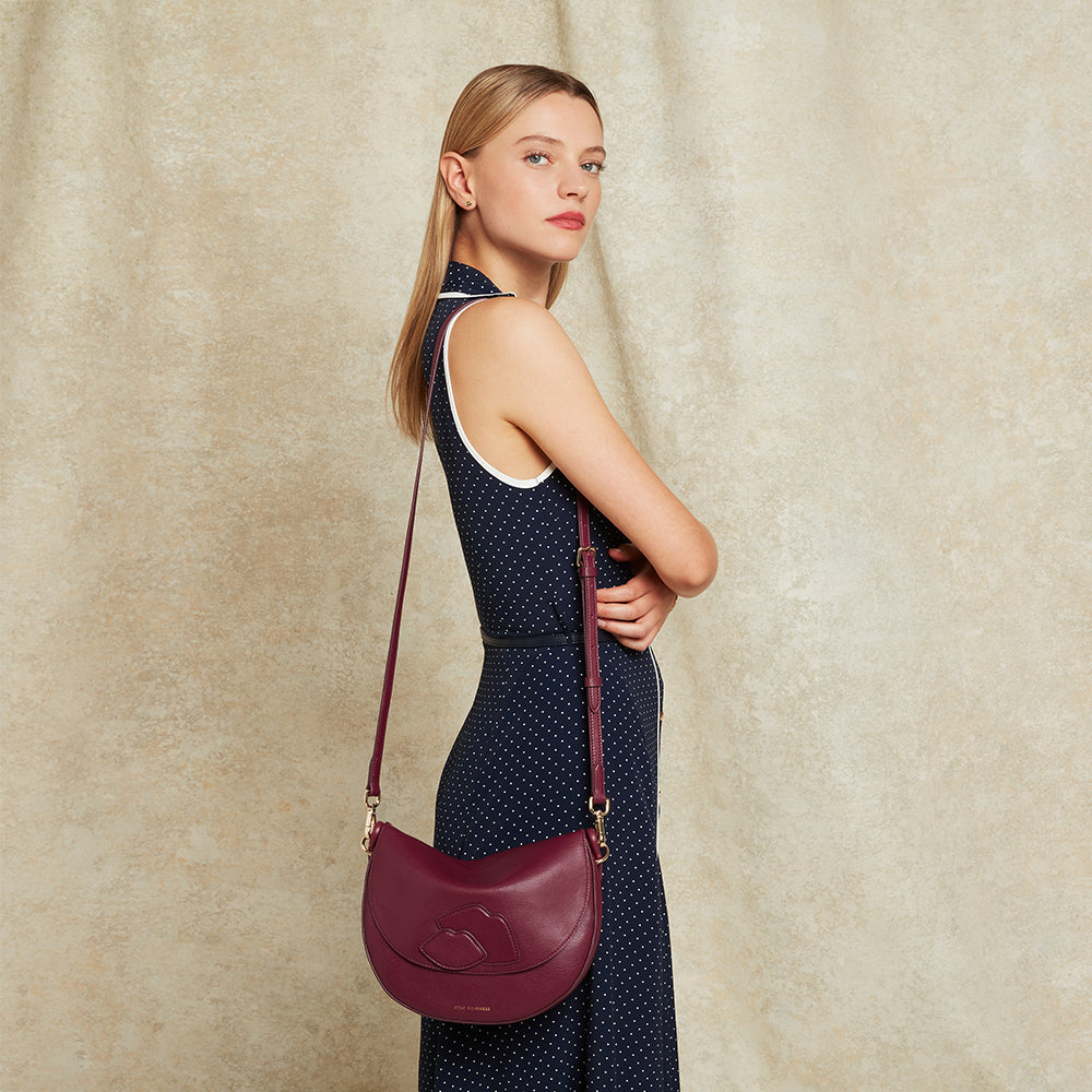 Woman in a navy polka dot dress wearing a burgundy leather bag showing the crossbody strap and lip details against a beige background.