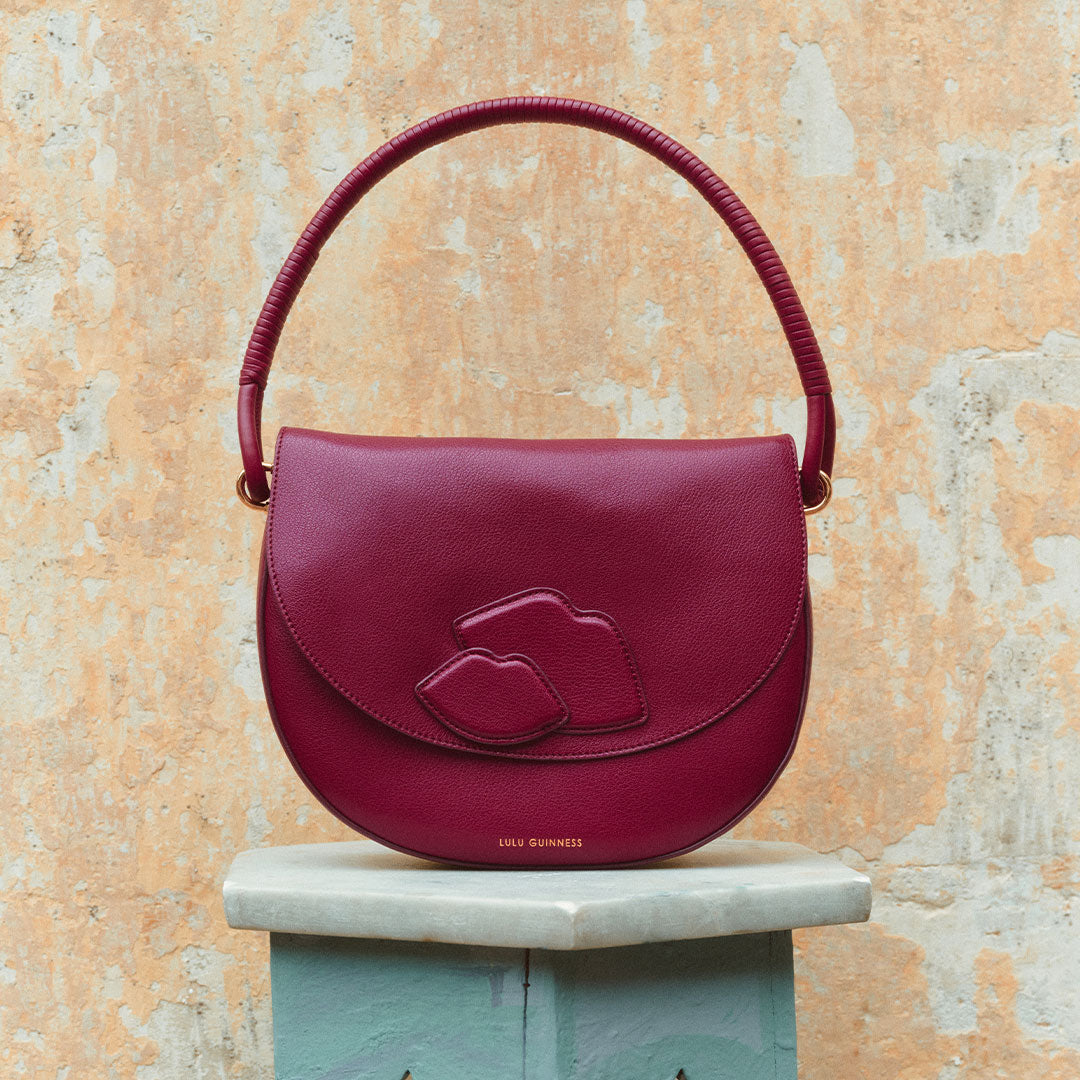 Burgundy handbag with lip design on a marble pedestal against a textured wall.