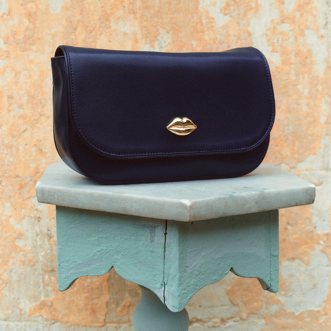 Navy blue handbag with a gold lip-shaped clasp on a decorative stand against an orange and beige textured wall.