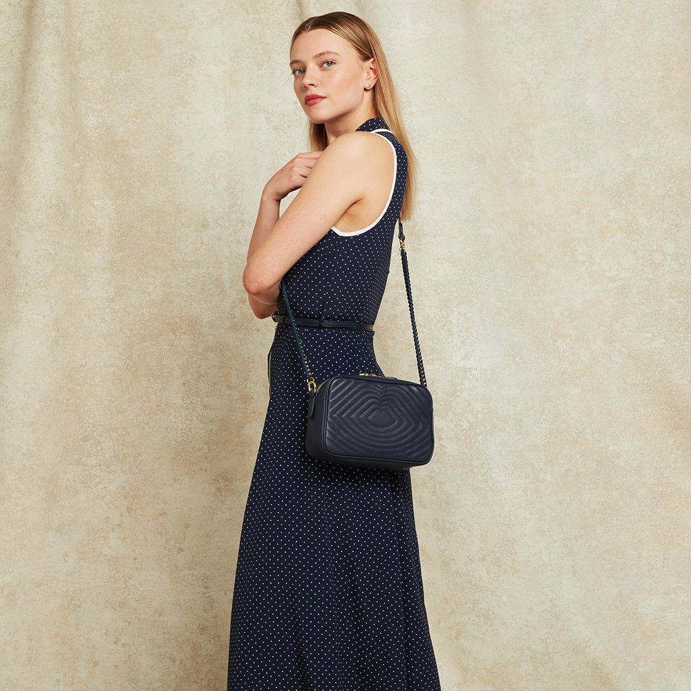 Woman in a navy polka dot dress holding a navy leather quilted crossbody bag against a beige background