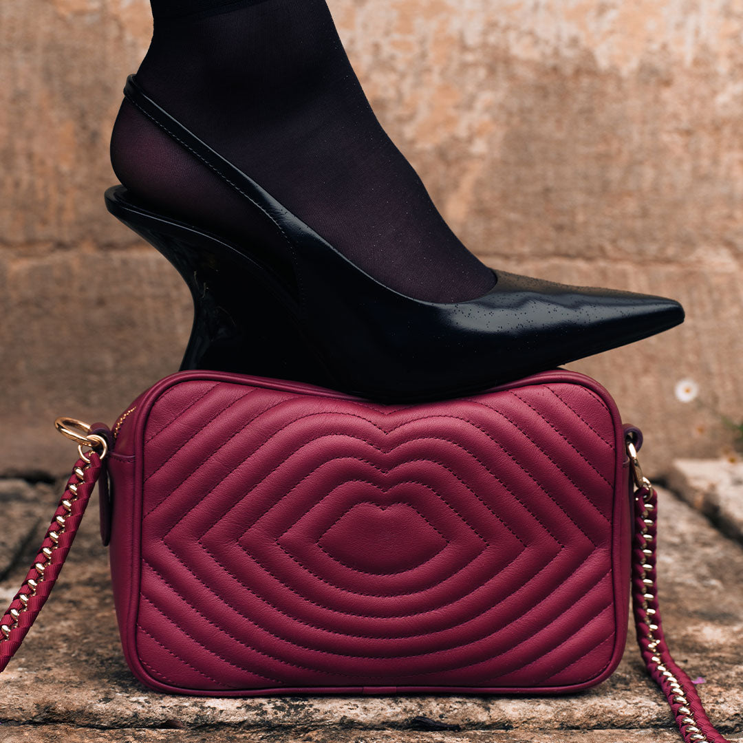Burgundy red quilted leather handbag with gold chain strap on stone surface, black high heel shoe on top.