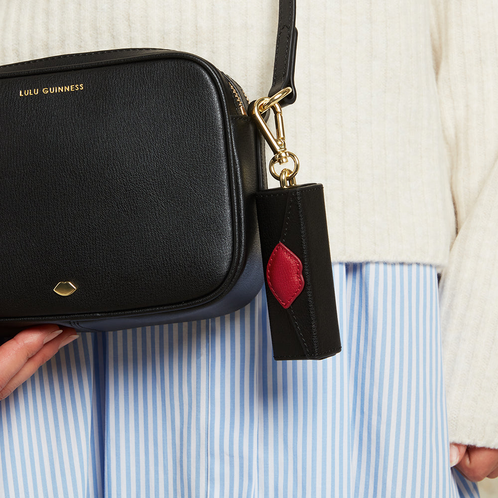 A black and red leather lip key charm clipped onto a black bag and held by a person wearing a white sweater and blue striped shirt.