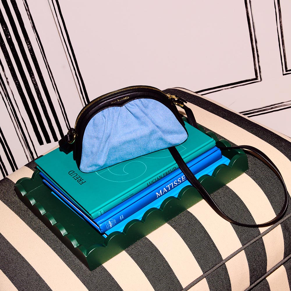 A denim and black leather crossbody bag sitting on top of a pile of books on a black and white striped chair.