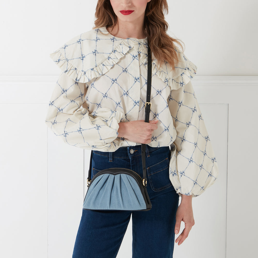 A woman in a cream blouse with blue bows and dark-wash jeans is modeling a half-moon shaped denim crossbody bag with black leather trim.