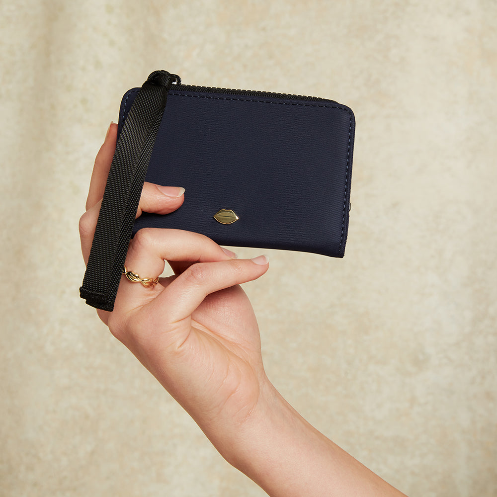 Hand holding a navy blue wallet with a black ribbon zip pull against a beige background
