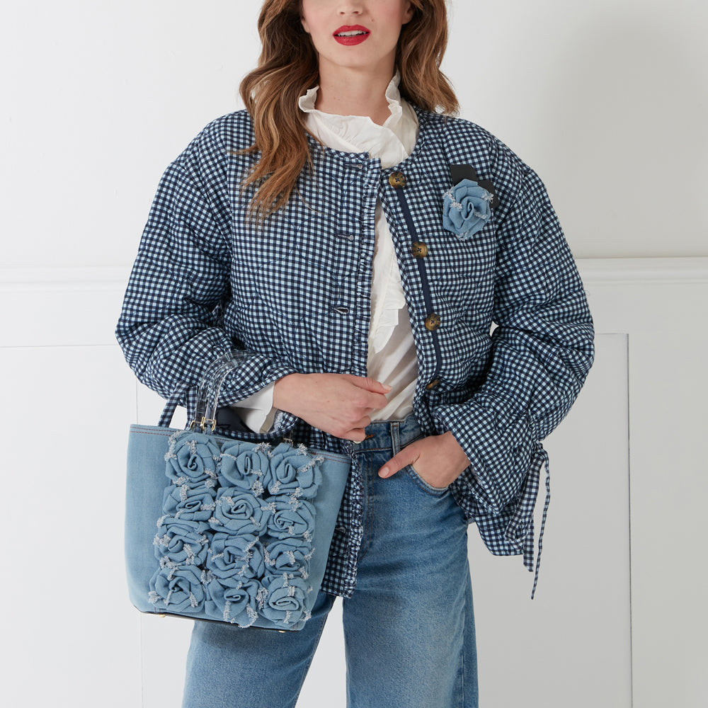 A woman in a blue gingham quilted jacket and jeans holds a light blue denim handbag adorned with fabric roses. The background is white.
