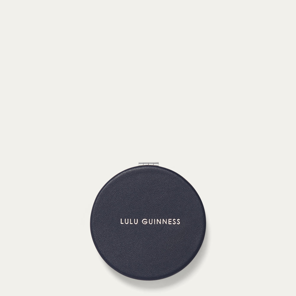 Round navy compact mirror with 'Lulu Guinness' branding on a light gray background