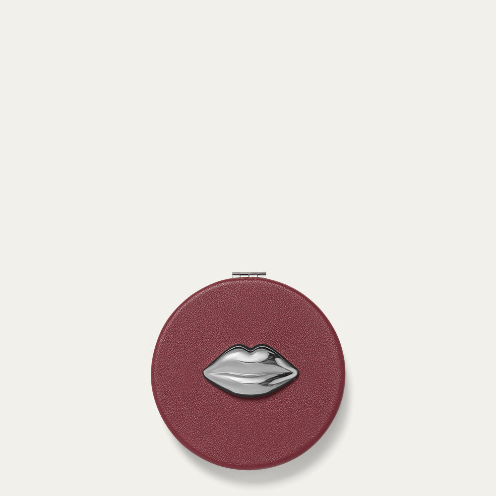 Round burgundy red compact mirror with a silver lip design on a light beige background