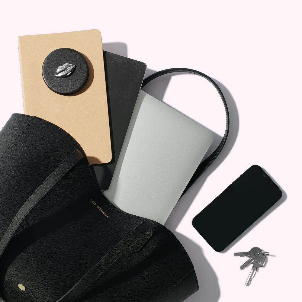 A black leather tote bag handbag with a laptop, notebooks, smartphone, keys and compact mirror spilling out.