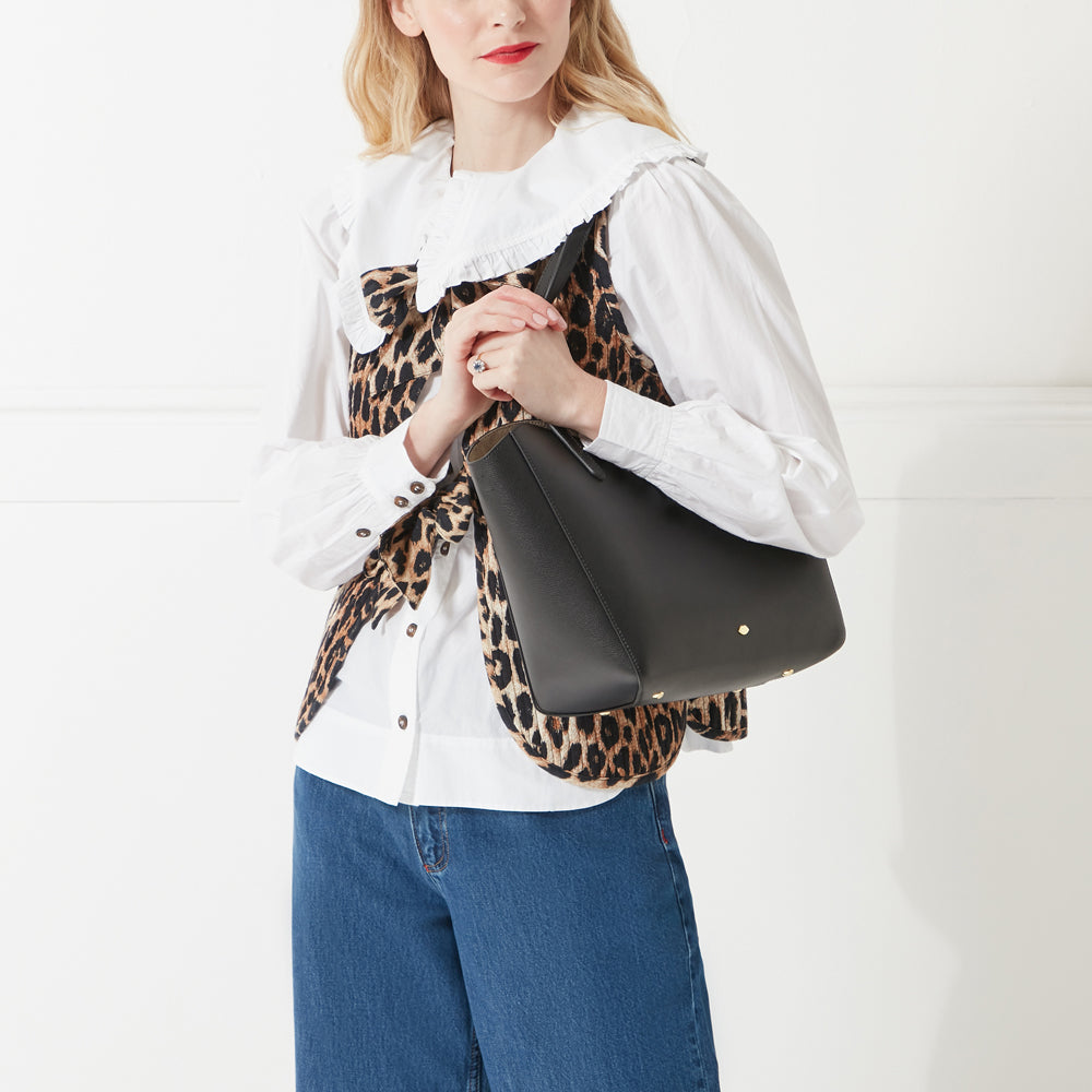 Model wearing a white shirt, jeans and a leopard print waistcoat holding a black leather tote bag with lip stud detail on her shoulder.