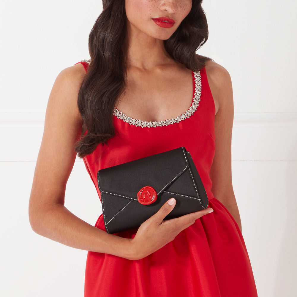 Woman in a red dress holding a black envelope clutch with a red wax seal.