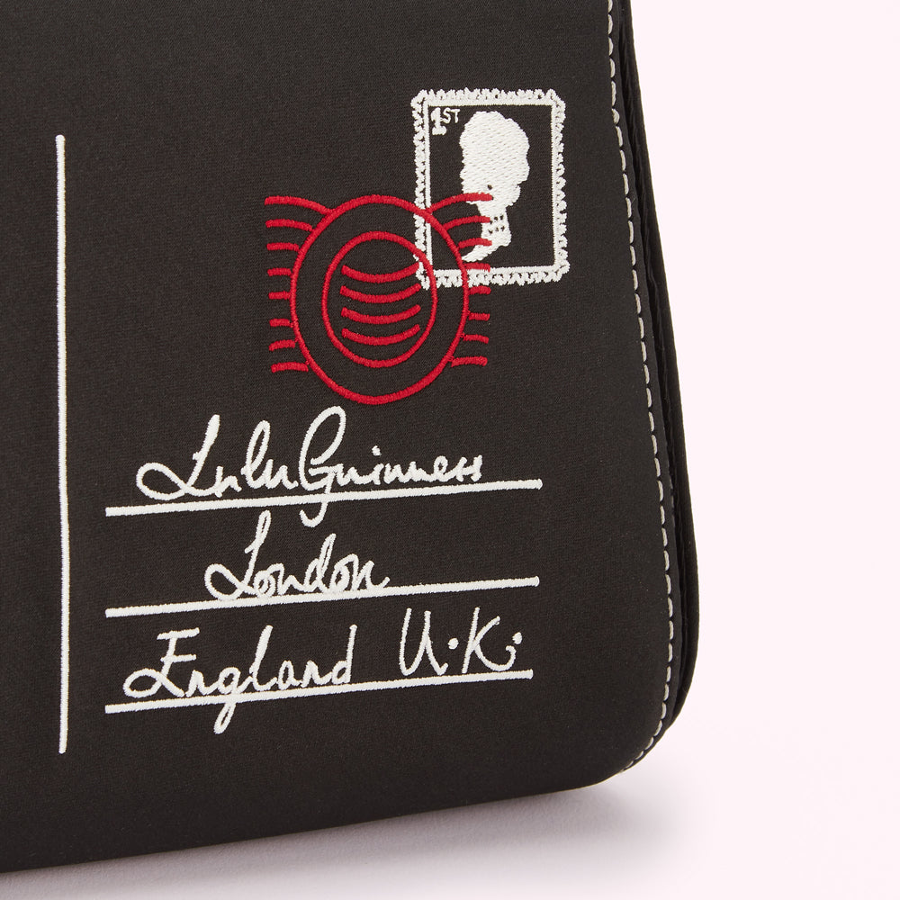 Close-up of a black accessory with embroidered details resembling a postage stamp and address, including the name "Lulu Guinness," "London," and "England U.K." in red and white thread.