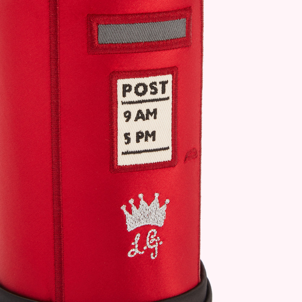 Close-up of a red fabric postbox with embroidered details, including a crown and the letters "L.G.", against a white background.
