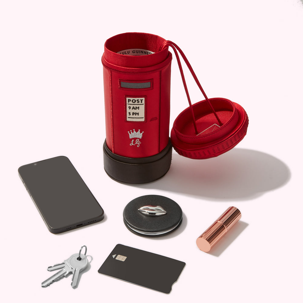 Red postbox-shaped handbag with its lid open, next to a smartphone, keys, a credit card, and lipstick.