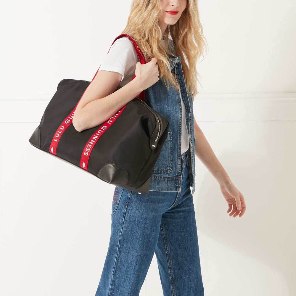 Model wearing a denim outfit, white trainers and red lipstick wearing a black nylon holdall with red branded 'Lulu Guinness' handles on her shoulder.