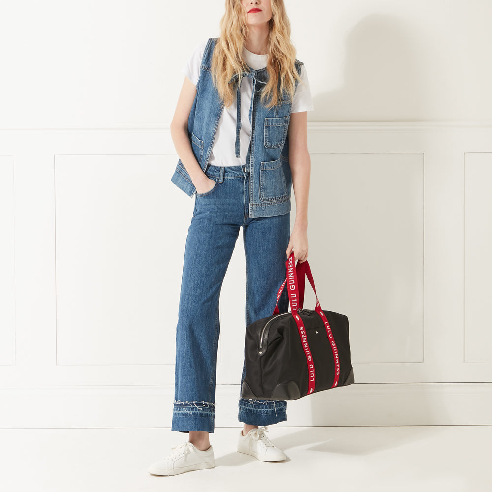 Model wearing a denim outfit, white trainers and red lipstick holding a black nylon holdall with red branded 'Lulu Guinness' handles.