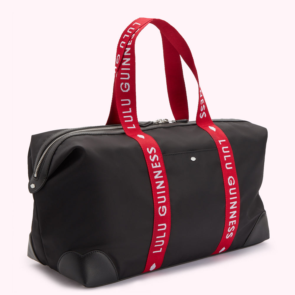 Side angle image of a black nylon holdall duffle bag with a red Lulu Guinness branded strap, silver metal lip pin on front pocket and curved leather corners.