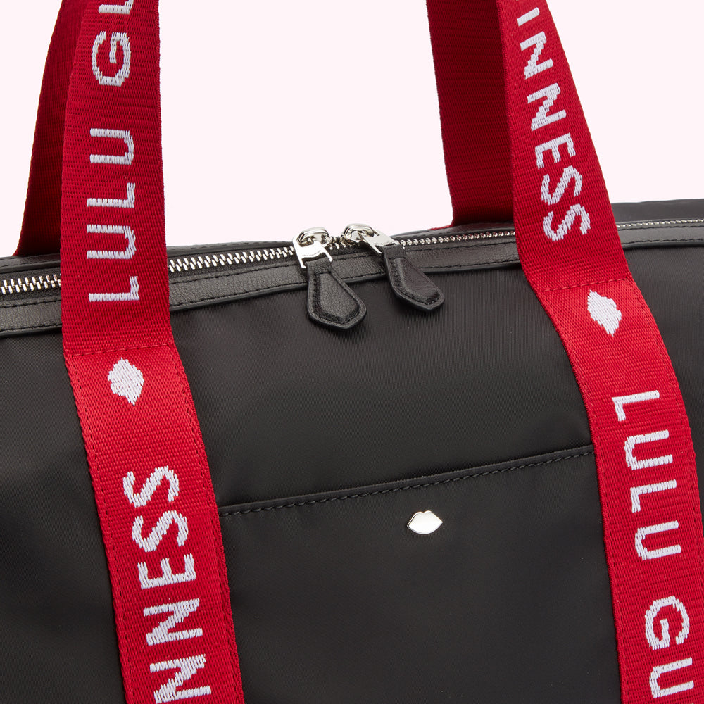 Close up detail of a black nylon holdall duffle bag with red Lulu Guinness branded straps, showing a silver lip pink detail on the front and silver metal zip detail.