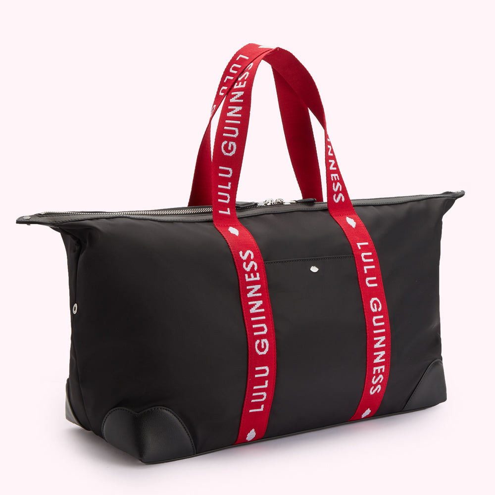 Side angle image of a black nylon holdall duffle bag with a red Lulu Guinness branded strap, silver metal lip pin on front pocket and curved leather corners.