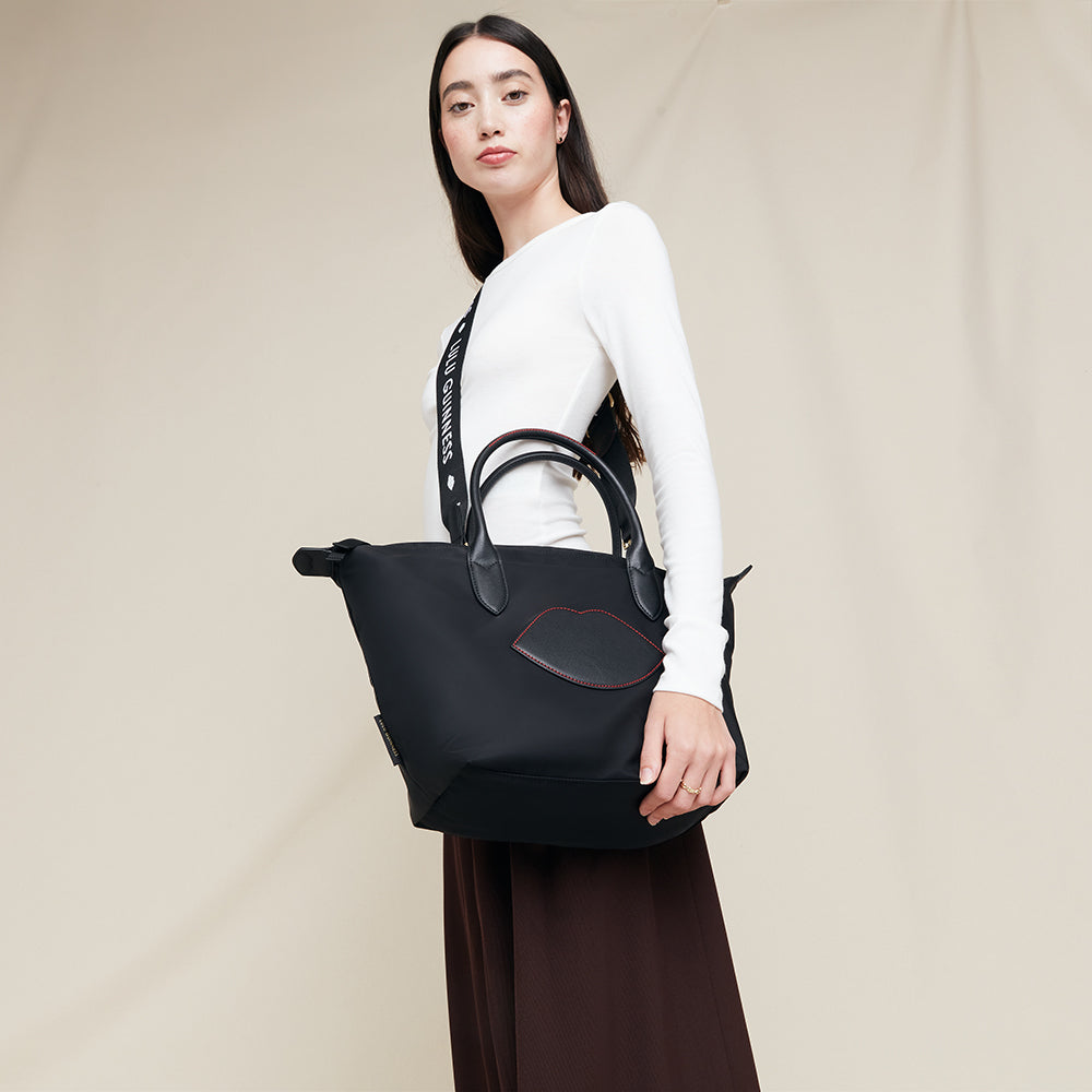 Woman holding a black tote bag with a detachable strap against a plain background