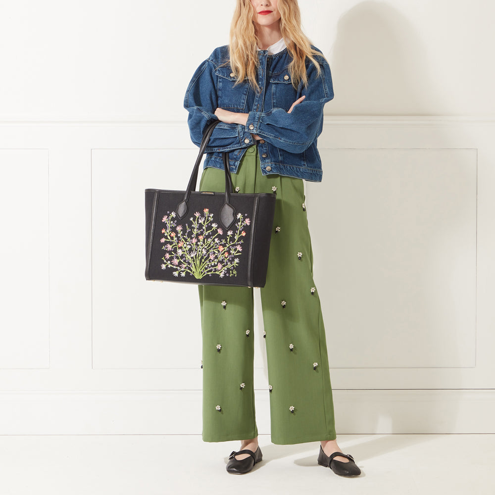 Model wearing a denim jacket, green trousers and red lipstick holding a black canvas and leather floral embroidered tote bag.