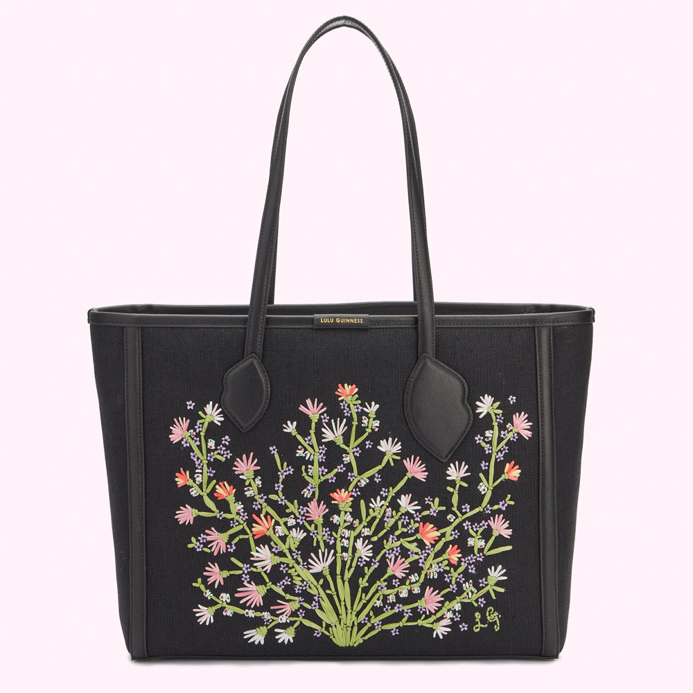 Front image of a black canvas and leather tote bag showing pastel floral embroidery, gold foil print Lulu Guinness logo on top edge and lip shaped handle anchors.