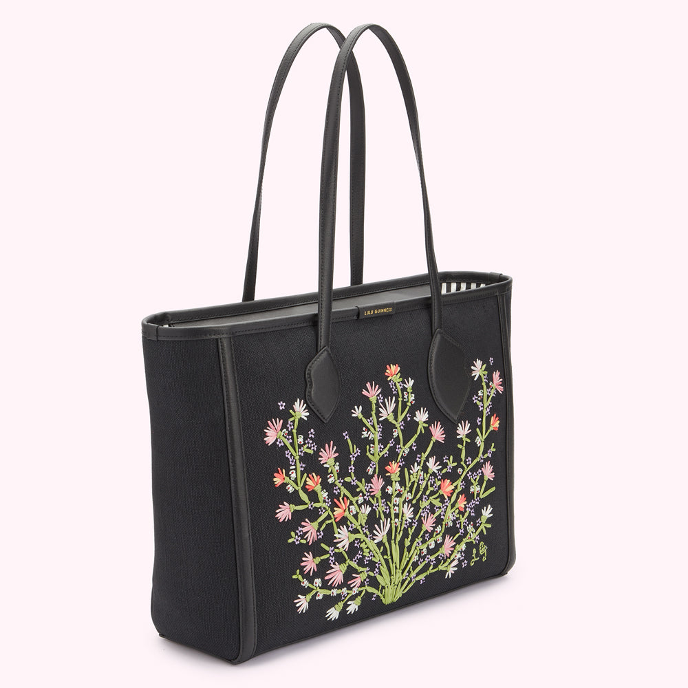 Angle side image of a black canvas and leather tote bag showing pastel floral embroidery, gold foil print Lulu Guinness logo on top edge and lip shaped handle anchors.