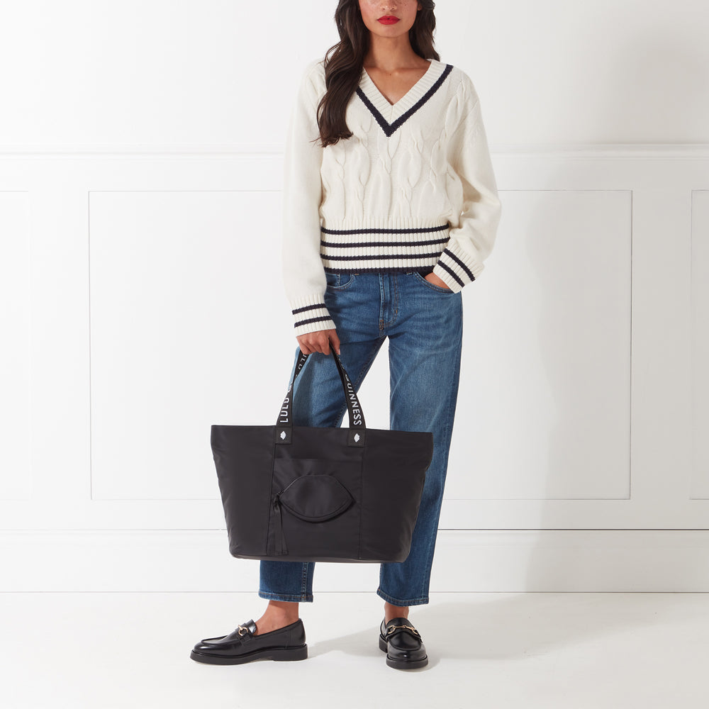 A woman in a cream V-neck sweater, blue jeans, and black loafers holds a large black tote bag with a small zippered pocket on the front. The bag has white lettering on the straps. The background is white.
