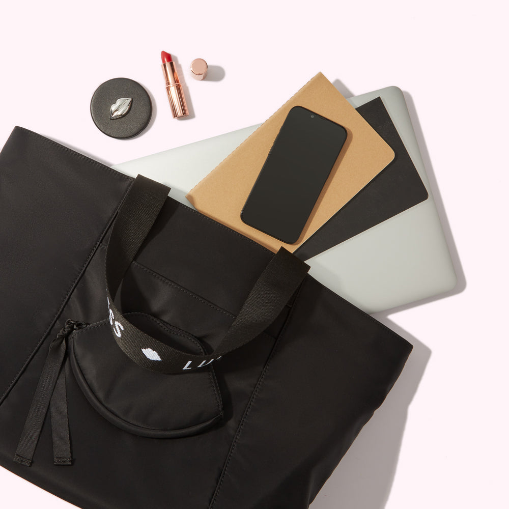 A black tote bag with a small black zippered pouch on the front containing a laptop, notebook, smartphone and makeup.