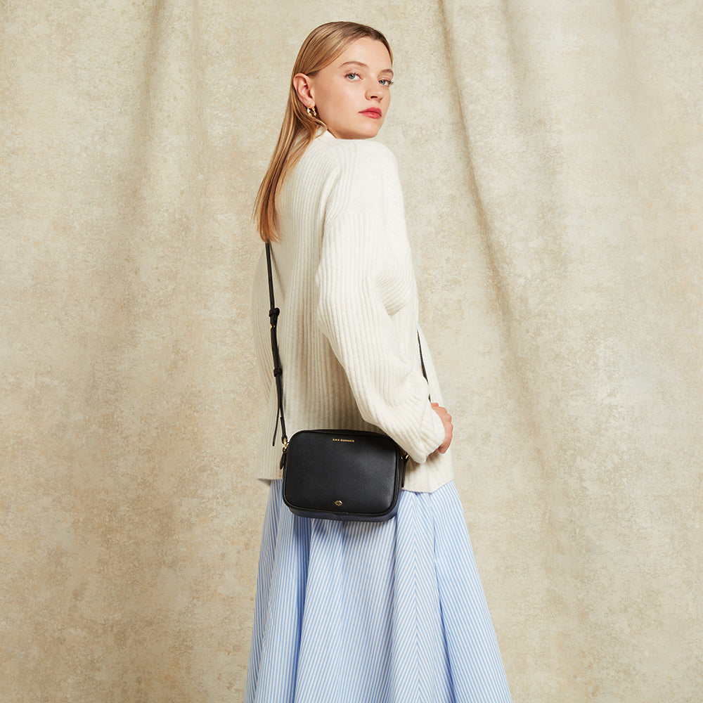 Woman wearing a black crossbody bag against a beige curtain background