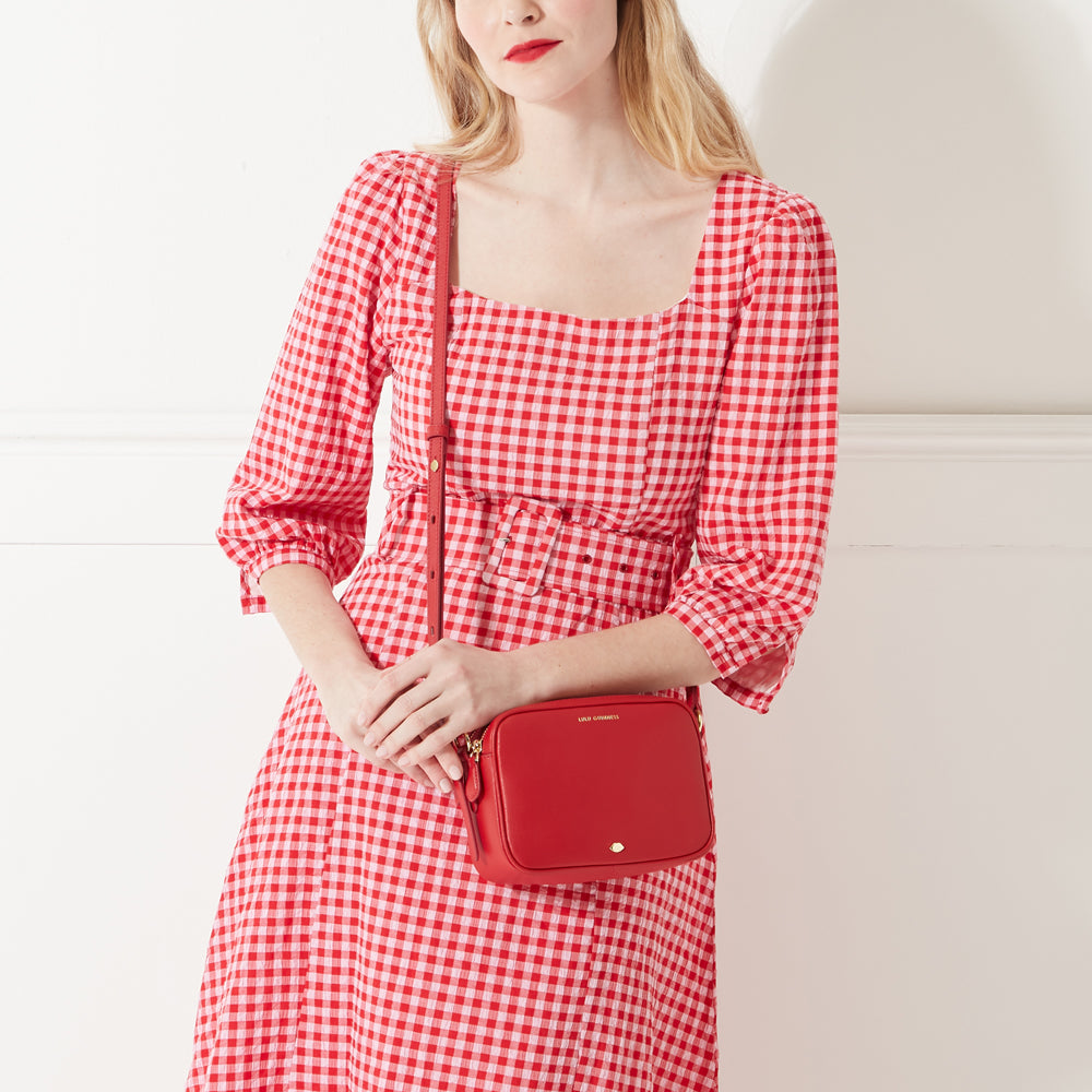 Woman with blonde hair modeling a red crossbody bag against a white background. She is wearing a red and white gingham dress.