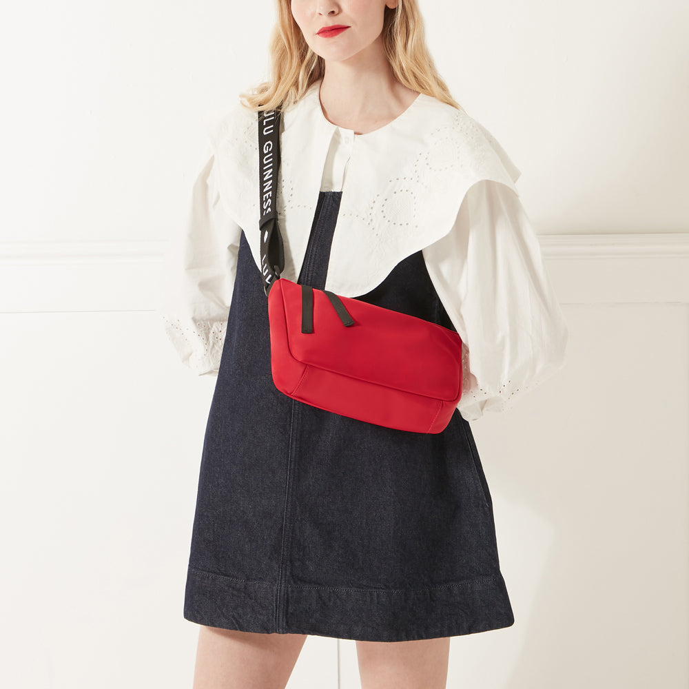 Model in a white shirt, denim dress and red lipstick wearing a red nylon shoulder bag with branded 'Lulu Guinness' shoulder strap.