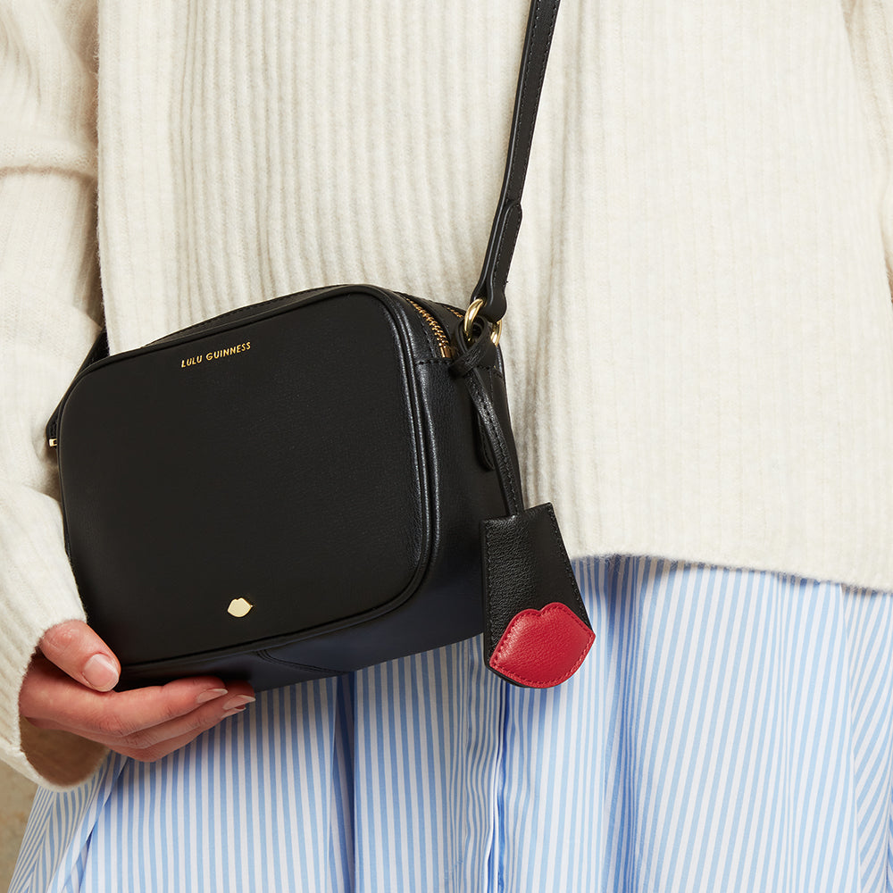 Black handbag with a red black with red lip charm held by a person wearing a white sweater and blue striped shirt.