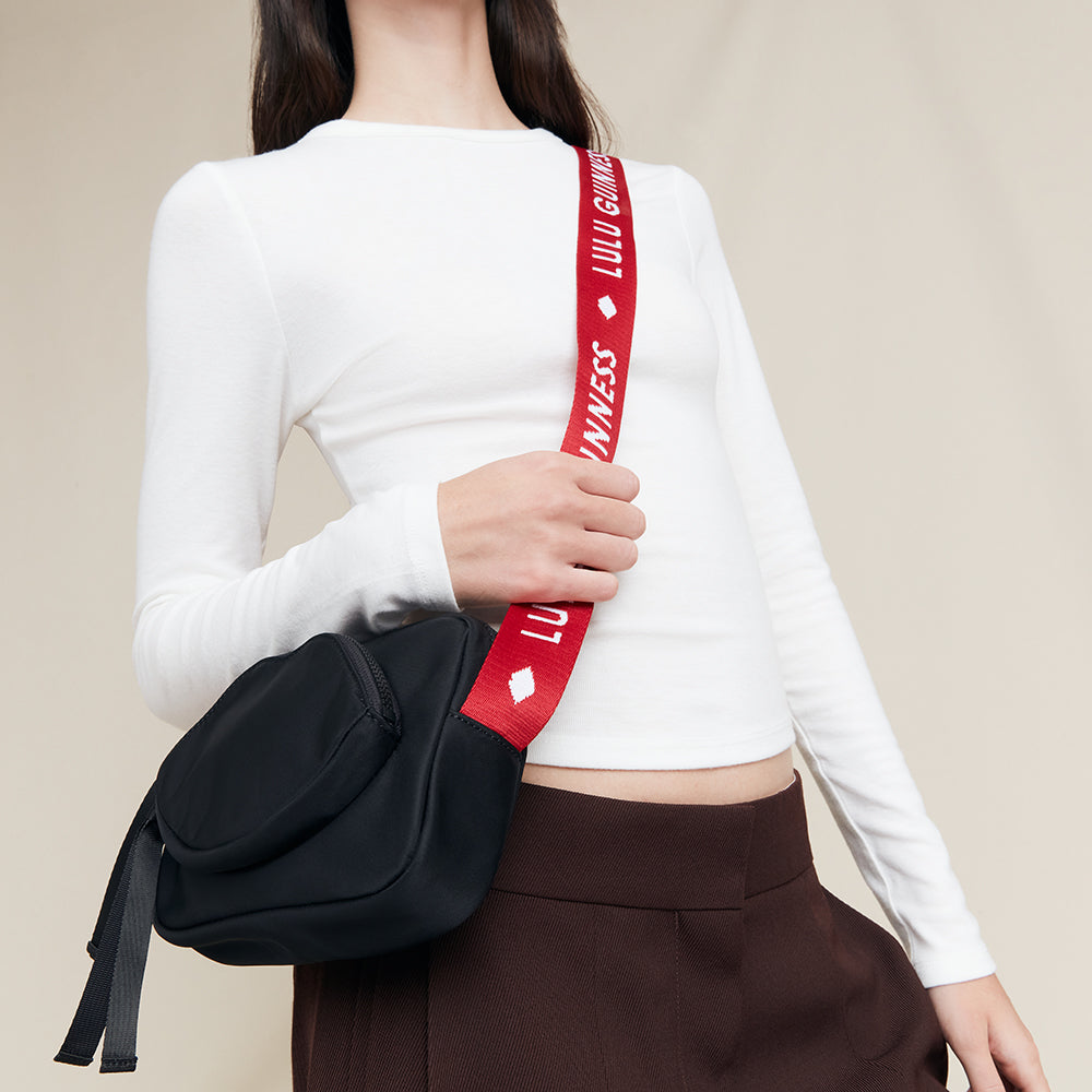 Person holding a black crossbody bag with a red strap featuring a 'Lulu Guinness' logo on a beige background