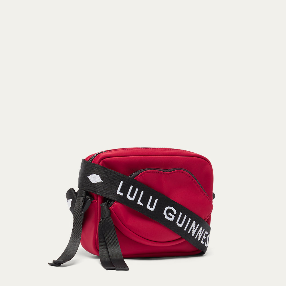 Red crossbody bag with a black strap featuring 'Lulu Guinness' and a lip-shaped pocket on a light gray background