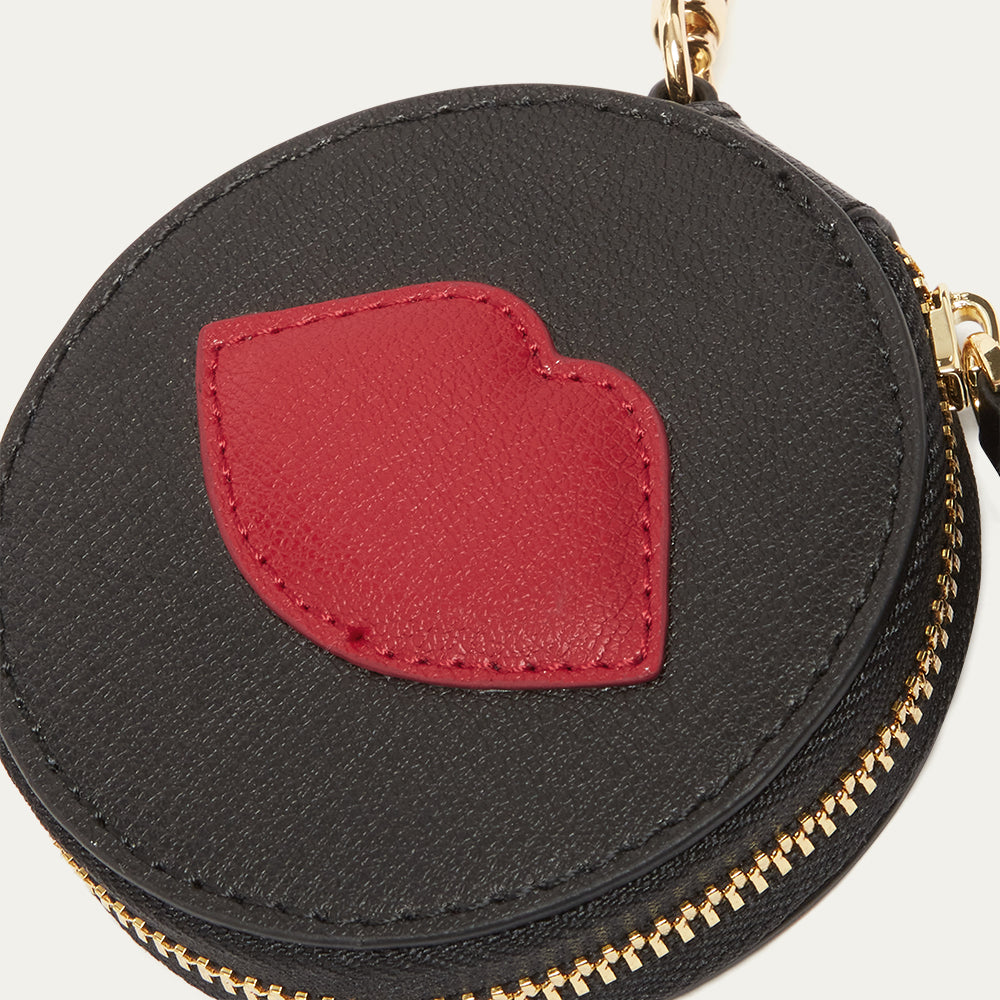 Black round pouch charm with a red lip-shaped patch and gold zipper on a light gray background