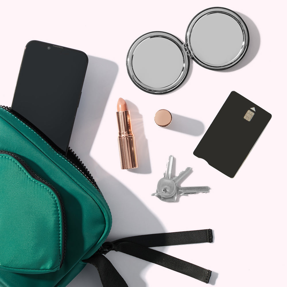 A green crossbody bag spills its contents: keys, a compact mirror, lipstick, compact mirror and a smartphone. All items are arranged on a white background.