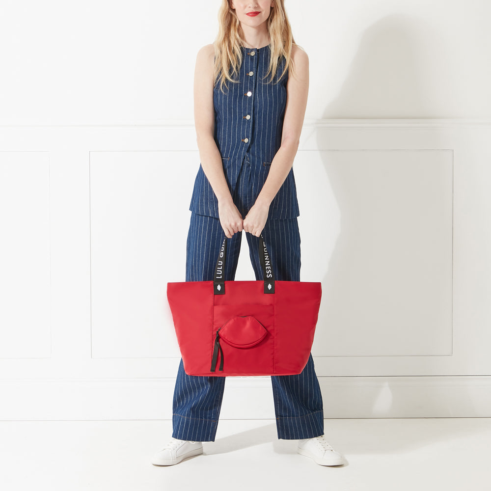 Model in denim outfit holding a red nylon tote bag with lip shaped pocket and branded 'Lulu Guinness' straps.