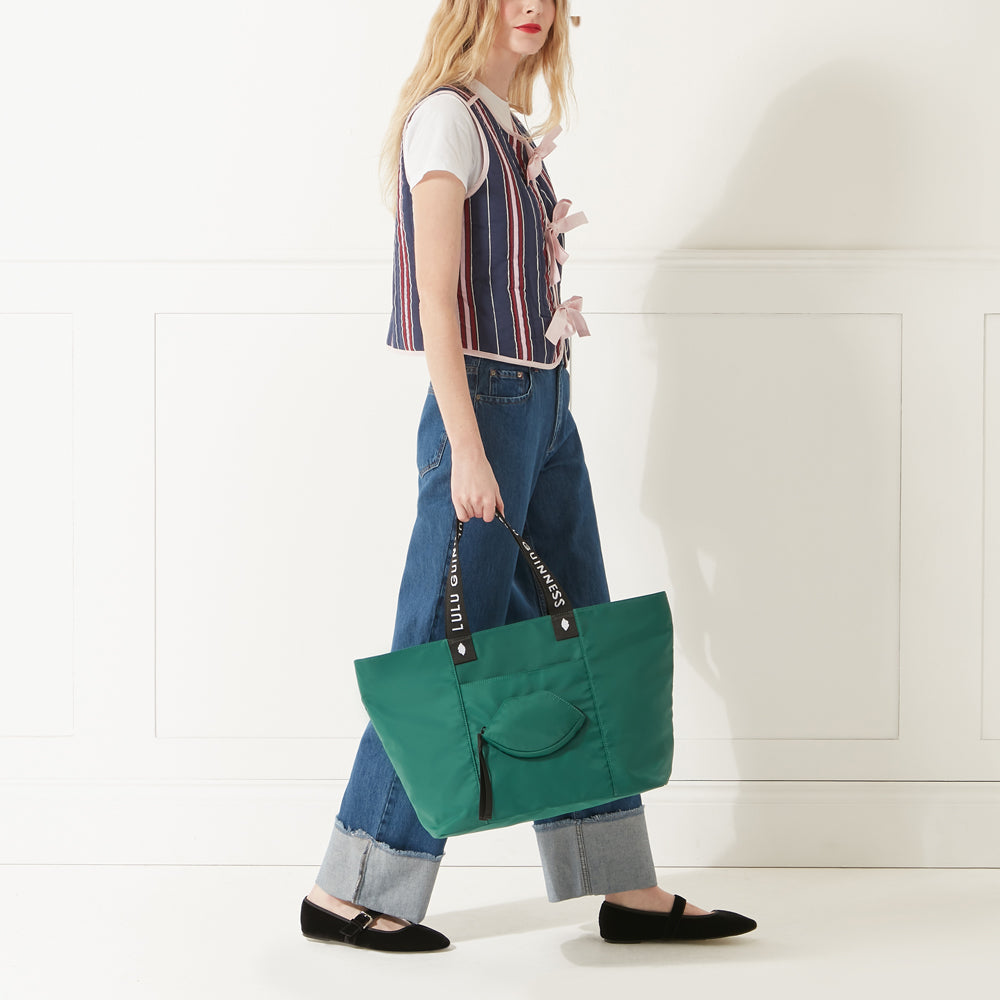 Model in a stripe gilet and denim jeans holding a green nylon tote bag with lip shaped pocket and branded 'Lulu Guinness' straps.