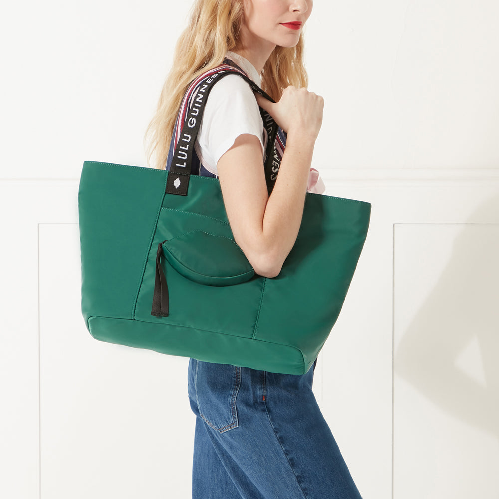 Model in a stripe gilet and denim jeans wearing a green nylon tote bag with lip shaped pocket and branded 'Lulu Guinness' straps on her shoulder.